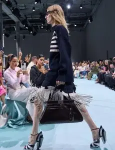 A model strutting down the fashion show runway, carrying a sleek black handbag and wearing a stylish outfit with fringe details. Audience members in the background are capturing the moment with their phones.